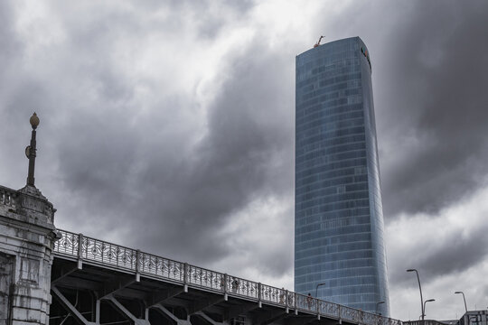 Bilbao, Pais Vasco, Espa&ntilde;a. 1 de Serptiembre 2026. Vista contrapicada de la imponente Torre Iberdrola alz&aacute;ndose junto a la balaustrada del puente de Deusto bajo un cielo tormentoso.
