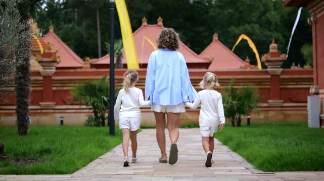 Three people continue family walk through tropical garden, with tall adult centered between two small children and hands linked. Pathway, grass, and temple style architecture form peaceful backdrop