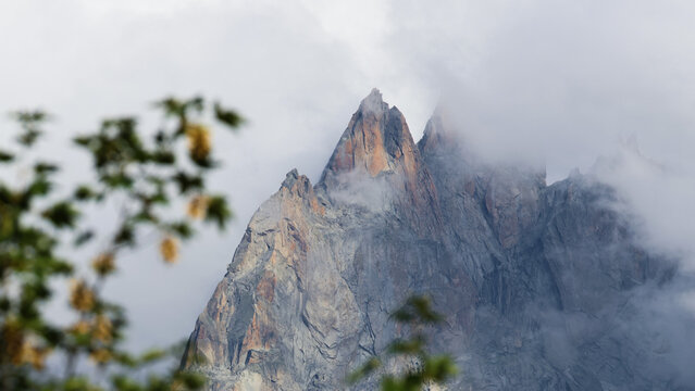 View of the rugged peaks of the Mont Blanc massif partially obscured by clouds with blurred foliage in the foreground in Chamonix, Auvergne-Rhone-Alpes, France.