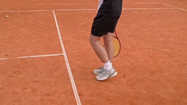 Caucasian young adult man walking on outdoor clay court holding tennis racket and bouncing ball. Footage serves commercial sports advertising, athletic training campaigns, fitness promotions