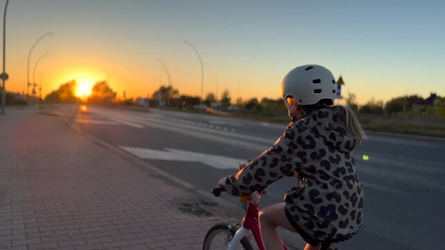 Young cyclist riding bicycle beside broad road at sunset, helmet on and hands on handlebars. Golden light glows behind rider, and calm evening setting gives simple commute feeling with open space.