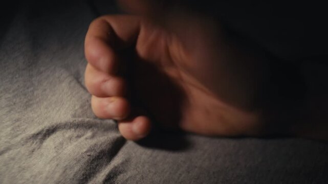 Male hand on a bed sheet slowly curls from open palm into a tense fist in dim moody light.