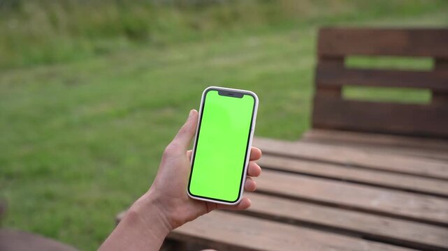 Hand holds smartphone with green screen outdoors. Phone centered in frame. Background shows wooden bench and grassy area. Person appears calm and focused. Ideal for app, social media