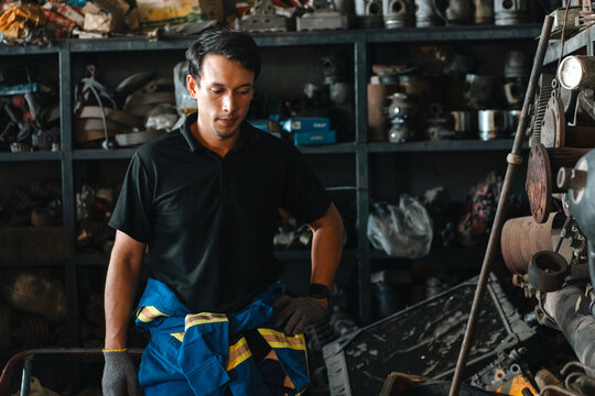 Electric vehicle battery car service Male worker wearing protective clothing in industrial workshop, surrounded by tools, equipment, and metal parts with focused expression and subdued light