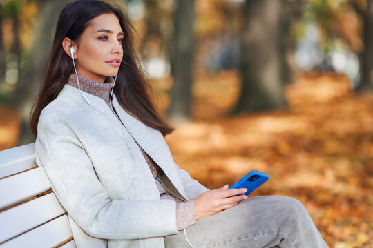 A young beautiful woman is sitting on a bench in an autumn park and talking on the phone. A girl listens to music in the autumn forest