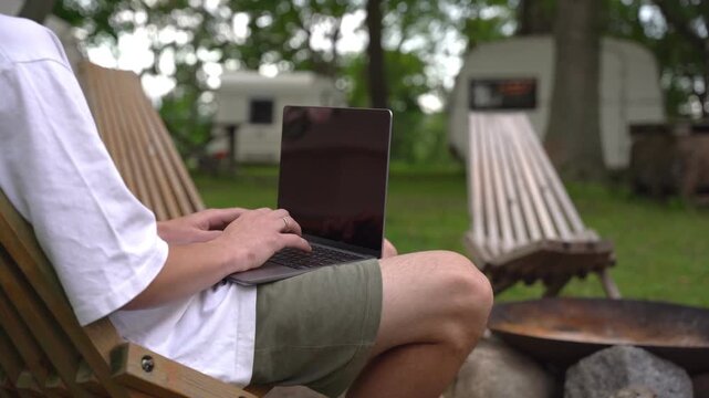 Person sits relaxed in wooden chair, typing on laptop. Background shows campsite with trailers and trees. Scene conveys calm, productive outdoor work or travel vibe. Ideal for remote work, vacation