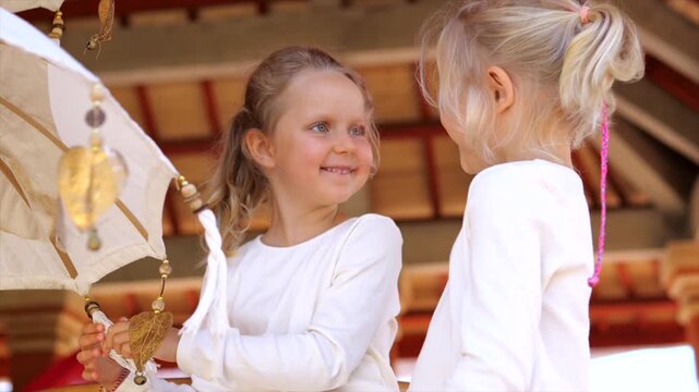 Two young girls handle hanging ornament together beneath parasol. Smiling children share focused play in warm wooden setting, passing tassels and decorative pieces between small hands during