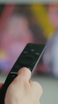 Vertical video. Woman hand switches TV remote control with finger, viewing of news, TV programs, modern technology for controlling equipment at a distance. Blurred TV in the background. Close up view.