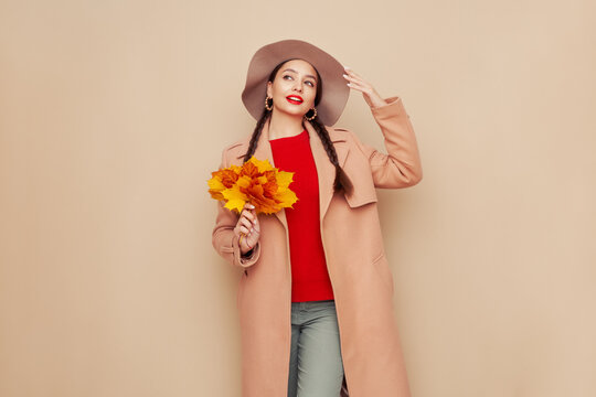 A beautiful young woman in an autumn fashionable outfit with shopping bags and round hat pointing aside at free space over beige background