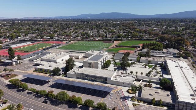 Orbiting aerial perspective of a modern high school campus in Santa Clara, California, featuring sports fields, solar panels on carports, and surrounding suburban neighborhoods on a clear day