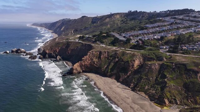 Aerial view of Mussel Rock Park and the Northern California coastline in Daly City, with ocean waves crashing on the sandy beach next to steep cliffs and a residential neighborhood