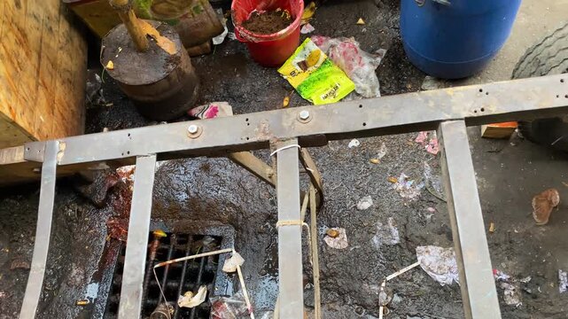 Metal handcart placed over dirty drainage manhole with debris, plastic barrel in dhaka city - top view.