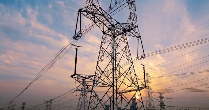 A high-voltage power transmission tower stands against a sunset sky with orange clouds, steel grid structures, and long cables.