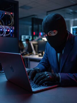 A man wearing a black balaclava and navy suit stares at the camera while holding an open laptop in a dark setting.