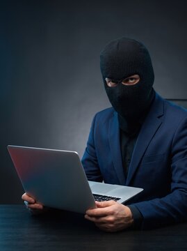 A man wearing a black balaclava and navy suit stares at the camera while holding an open laptop in a dark setting.