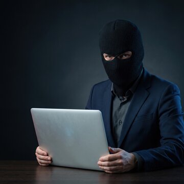 A man wearing a black balaclava and navy suit stares at the camera while holding an open laptop in a dark setting.