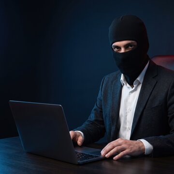 A man wearing a black balaclava and navy suit stares at the camera while holding an open laptop in a dark setting.