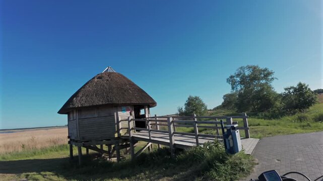 First-person chest-mounted view riding an electric bike behind cyclists on a cobbled section of the EuroVelo 10 route past a small thatched wooden house in Germany