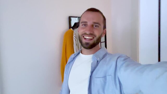 Young handsome man recording a video for his social media, showing his new modern home to his followers and waving at the camera while walking around and finally sitting on the sofa