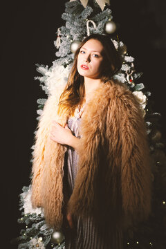 Elegant young woman in fluffy faux fur coat posing in front of a decorated Christmas tree