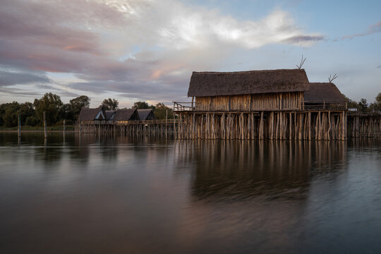Reconstructed prehistoric stilt houses glow in the golden light of sunset over the calm waters of Lake Constance.