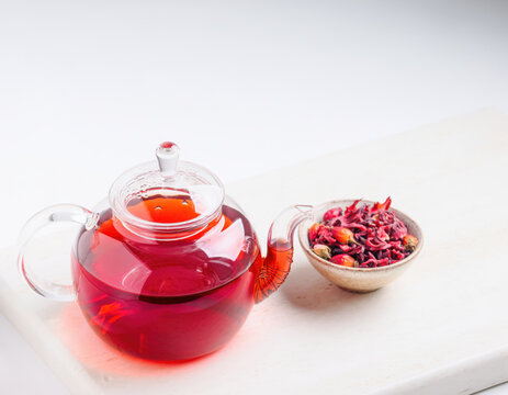 Clear glass teapot brimming with vibrant red hibiscus tea sits beside a small bowl of dried hibiscus flowers, ready for a healthy and refreshing herbal infusion on a clean white surface