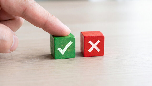 Human finger touches wooden green cube marked with white checkmark beside red square block displaying a bright cross symbol