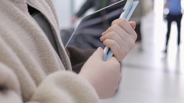 Close-up of hands holding a tablet in a busy indoor space. Person wears a beige fleece jacket. Background shows blurred people walking. Scene suggests modern public setting like airport or office