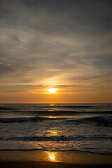 Obraz premium golden sunrise over sea horizon with wave reflections, vertical shot of ocean sunset with sun path on wet sand, scenic coastal sunrise with overcast sky and glowing water