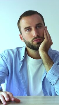 Young handsome man with a beard sitting at a table, looking sad and tired, showing boredom and disinterest. He rests his head on his hand, displaying signs of mental fatigue or depression