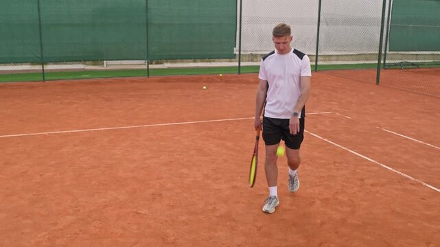 Caucasian young adult male tennis player walking on clay court and bouncing ball in slow motion tracking shot. Ideal for sports gear commercials or athletic fitness campaigns