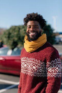 Smiling man in patterned red sweater with yellow scarf standing by car and christmas tree for seasonal holiday portrait and marketing use
