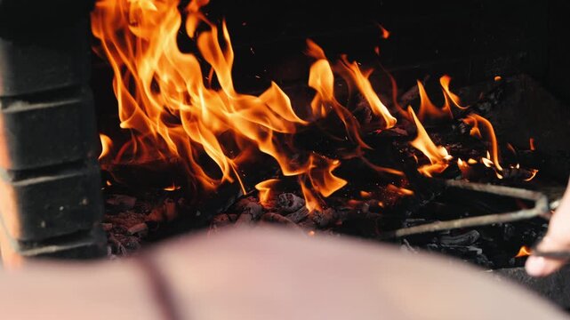 Over the shoulder view of a person stoking burning coals in a fireplace with a poker, bright flames rising in a dark stone hearth. High quality 4k footage