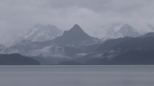 View of misty mountains rising above the calm water of Kachemak Bay under a heavy overcast sky in Homer, Alaska, United States.