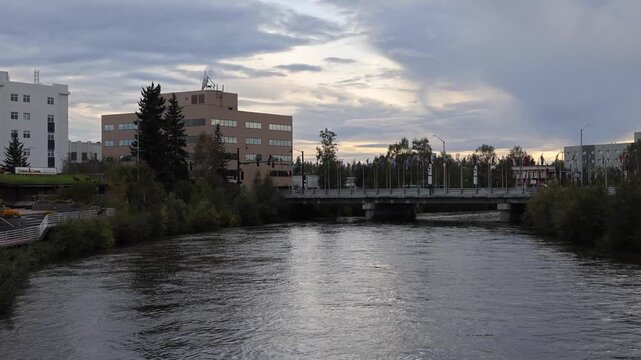View of the Chena River flowing under a bridge with urban buildings and trees under a cloudy sky in Fairbanks, Alaska, United States.