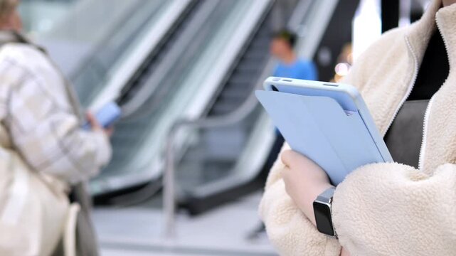 Person holds tablet while standing on escalator. Background shows blurred travelers moving. Focus remains on device and hands. Modern urban transit environment visible. Neutral tones dominate scene