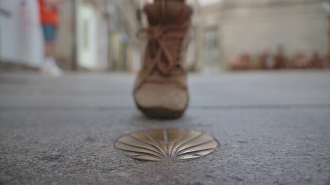 Close-up, low-angle perspective of a bronze scallop shell marker set into a stone pathway, with the out-of-focus legs and feet of a pedestrian walking in the background.