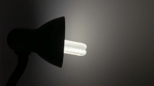 Close-up shot of a desk lamp with an energy-saving compact fluorescent (CFL) bulb turning on, instantly illuminating a pitch-black background