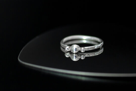 A silver ring with a central gemstone and two smaller stones on each side, placed on a reflective surface against a black background.