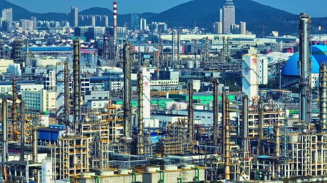 An aerial view of a large industrial refinery with tall distillation columns, complex piping, and a city skyline under bright sunlight.