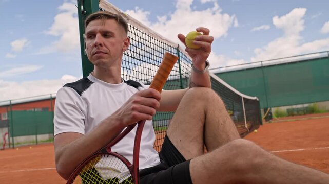 Caucasian young adult tennis player sitting on clay court holding racket and spinning ball during break. Ideal for sports commercial advertising and athletic training promotions