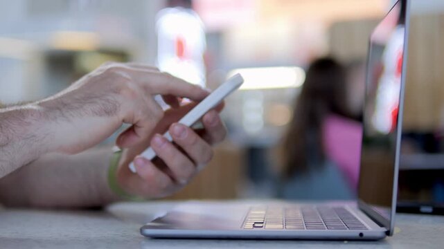 Hand uses smartphone while laptop sits nearby. Person focused, multitasking in casual setting. Perfect for scenes of digital work or social media use