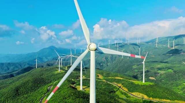 Aerial view of white wind turbines on green mountain ridges under bright sunlight with a blue sky and scattered white clouds.