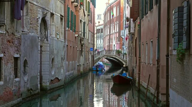 View of a narrow canal in Venice with historic brick buildings, a small stone bridge, and boats moored in the water in Venice, Veneto, Italy.
