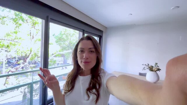 Joyful young woman recording a vlog or making a video call, excitedly showing off the keys to her new apartment while smiling and talking to the camera in her modern living room