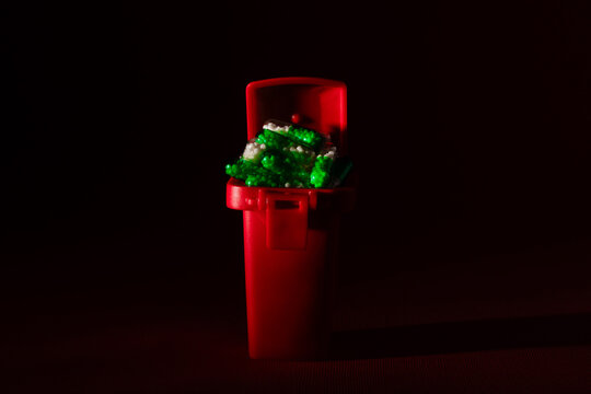 Small red trash bin filled with green and white plastic pieces, isolated on a dark background with dramatic lighting.