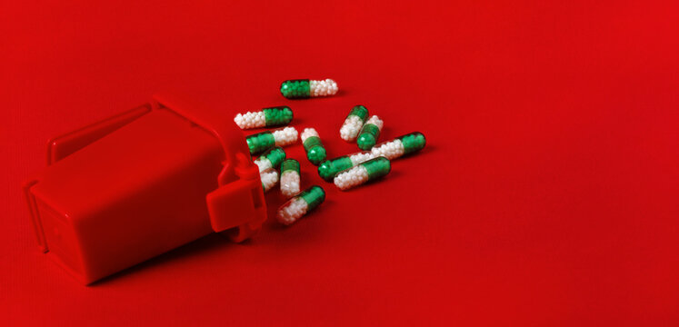 Green and white capsules scattered near a red medical container on bright red background, emphasizing healthcare, medication, and pharmaceutical themes.