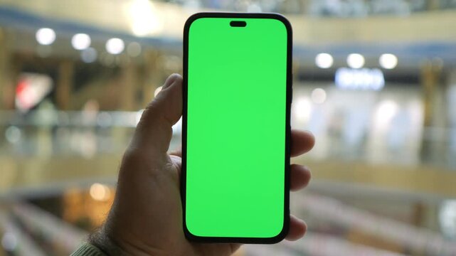 Hand holds smartphone with green screen. Blurred background suggests a public indoor space. No people visible in the scene. Use for showing digital content on screen