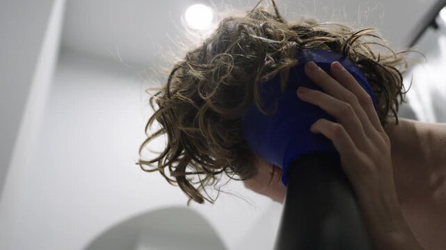 Close up of a woman using a hair dryer with a diffuser attachment to dry and shape her curls in the bathroom. The head is tilted while hands position the diffuser and encourage curl formation. Soft na