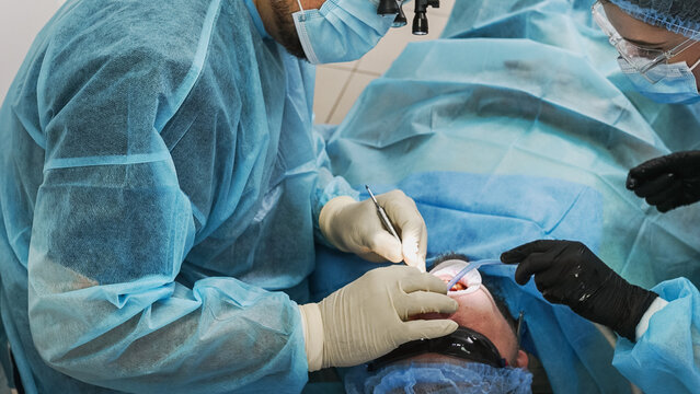 A male patient is undergoing dental treatment at a dental clinic; his mouth is open, a dental drill and saliva ejector are in use, and the dentist is working with an assistant.